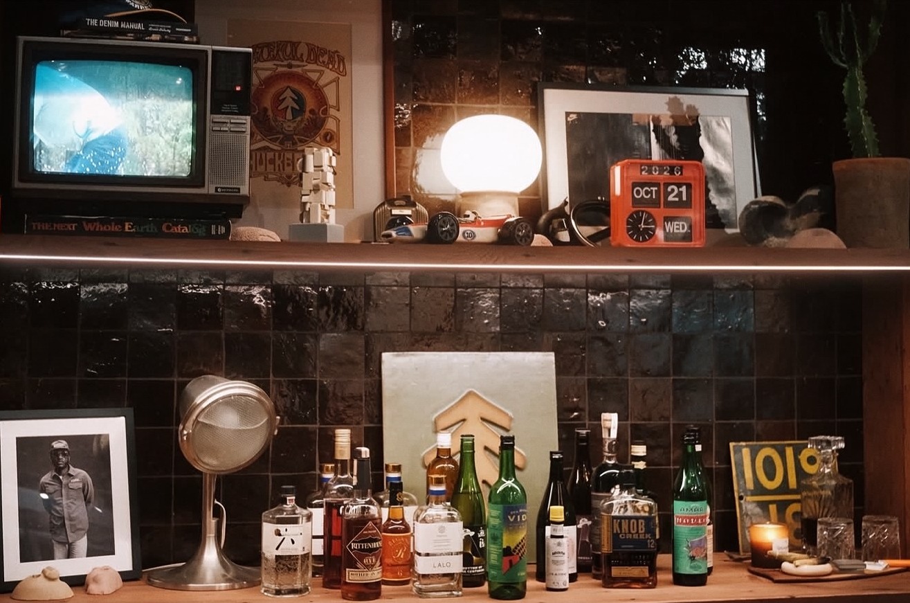 A bar's back shelves at night: a small CRT television, a glowing globe lamp and an orange flip clock above a dark tiled wall lined with bottles.