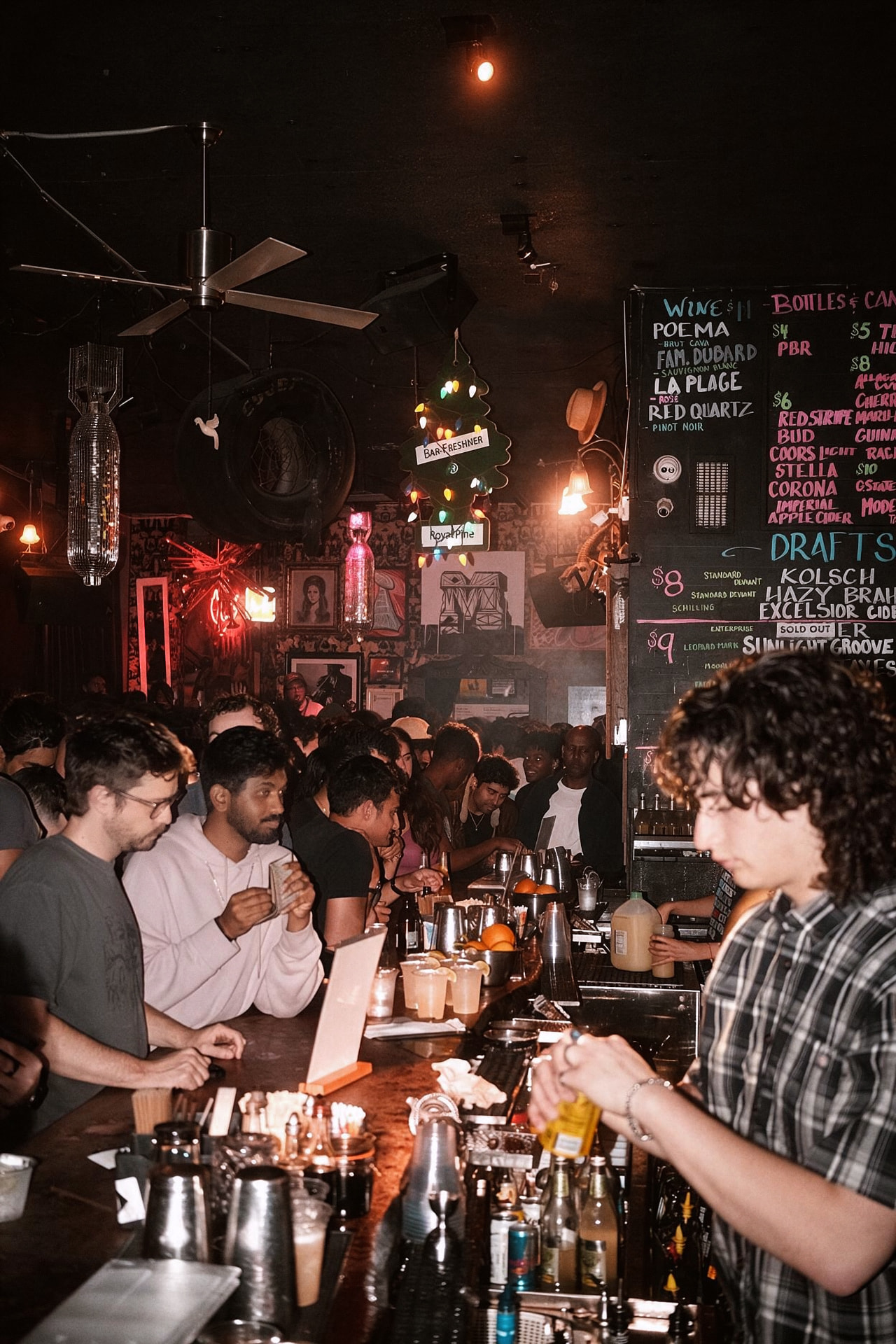 A crowded bar at night lit by flash and warm bulbs, a curly-haired bartender pouring in the foreground beneath a chalkboard drinks menu.