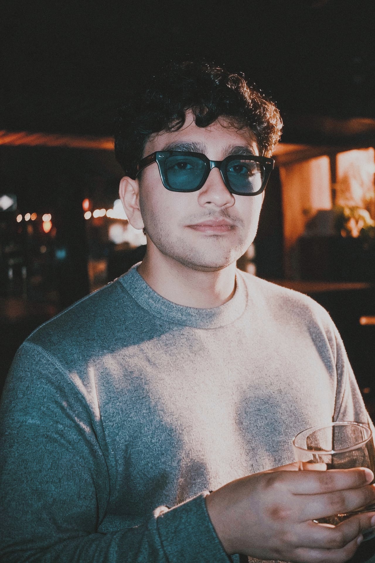 Flash-lit portrait of a curly-haired man in blue-tinted sunglasses and a grey sweater, holding a glass, warm bar lights glowing behind him.