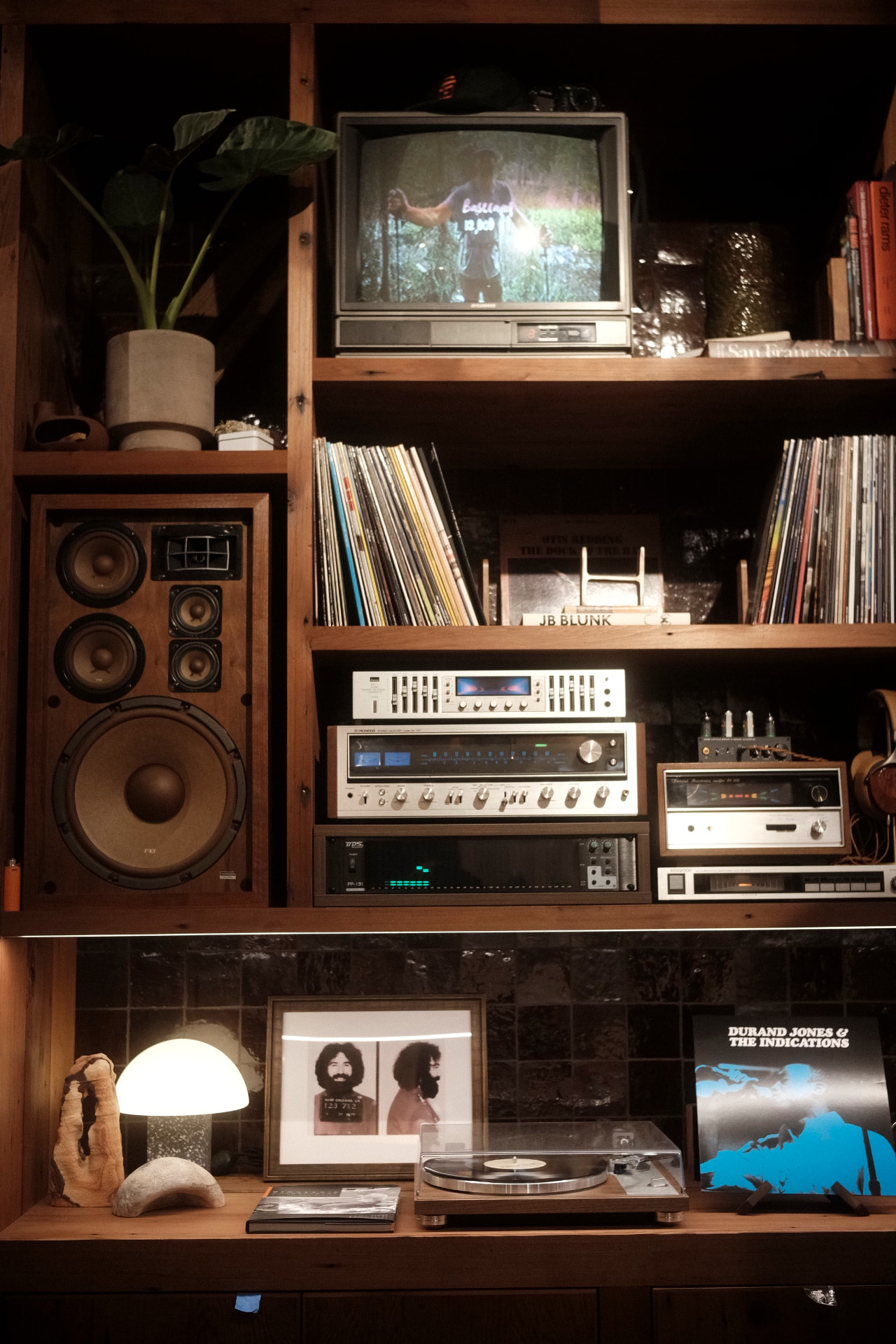Warm wooden shelving with a vintage speaker, stacked hi-fi receivers, a CRT television, records and a glowing mushroom lamp.