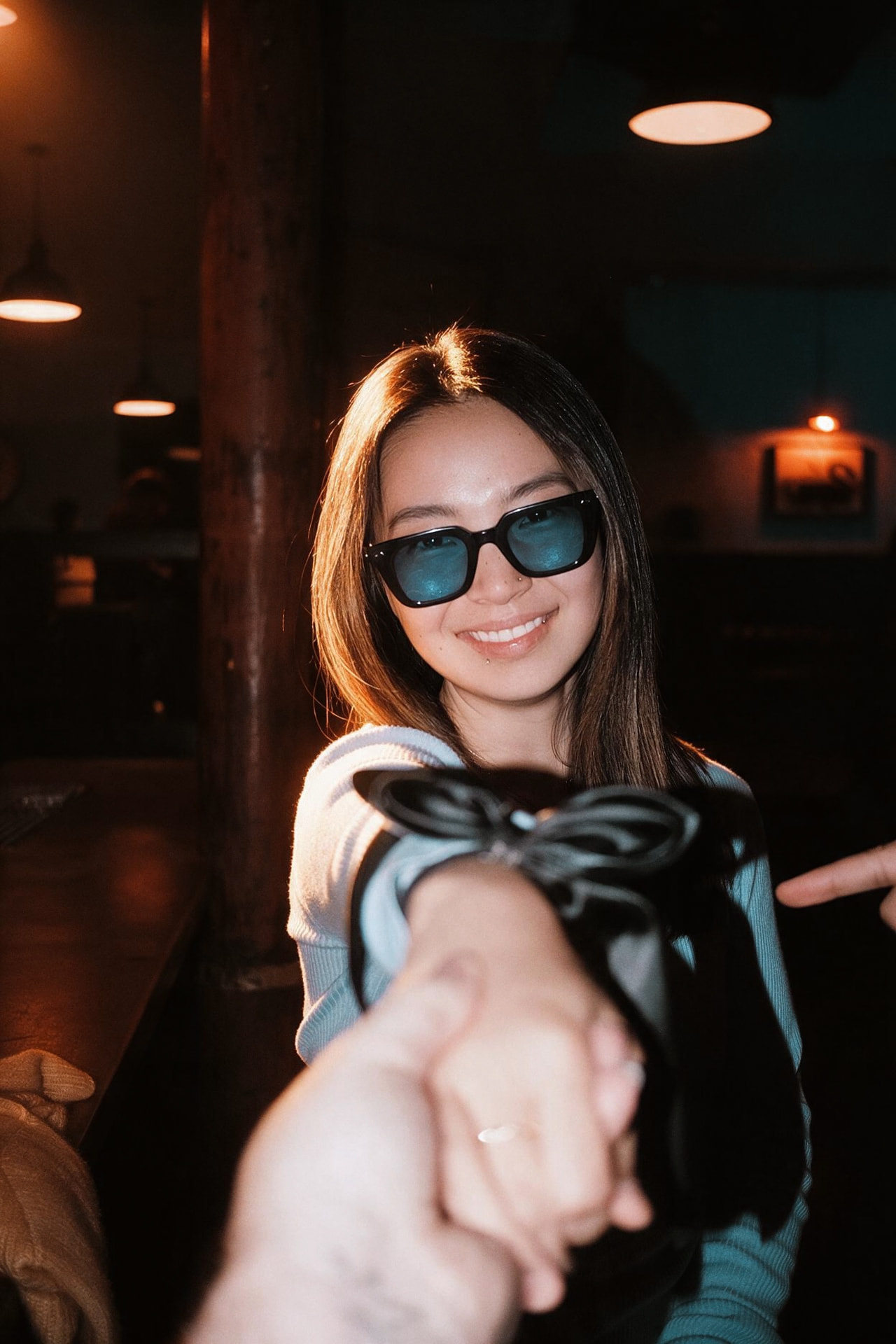 The same woman in blue-tinted sunglasses, smiling as she holds a hand reaching in from the camera, lit by flash beneath hanging lamps.
