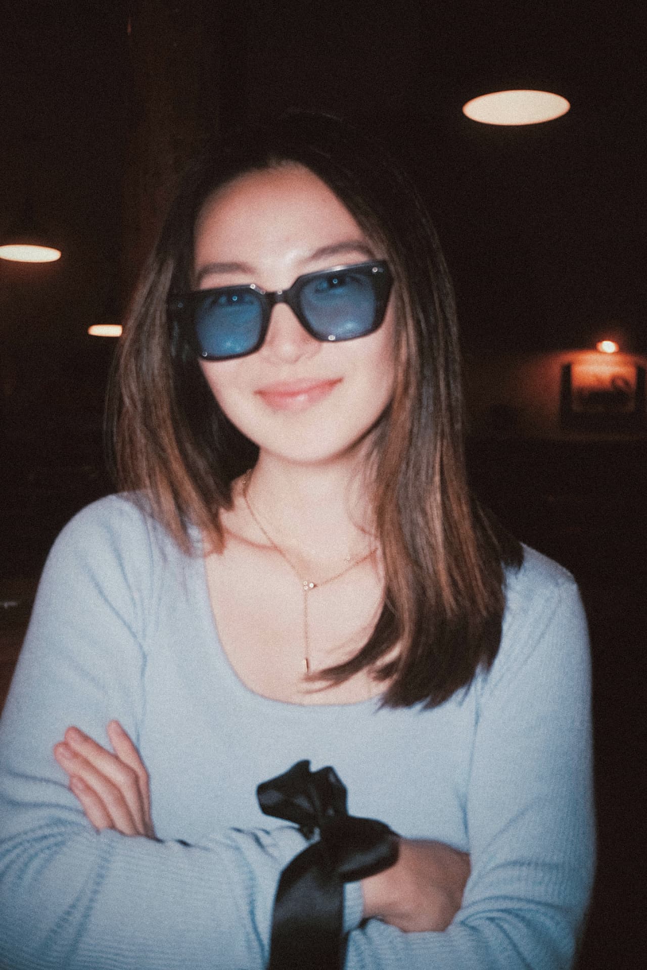 The same woman smiling in blue-tinted sunglasses, lit by flash against a dark room with round ceiling lights.