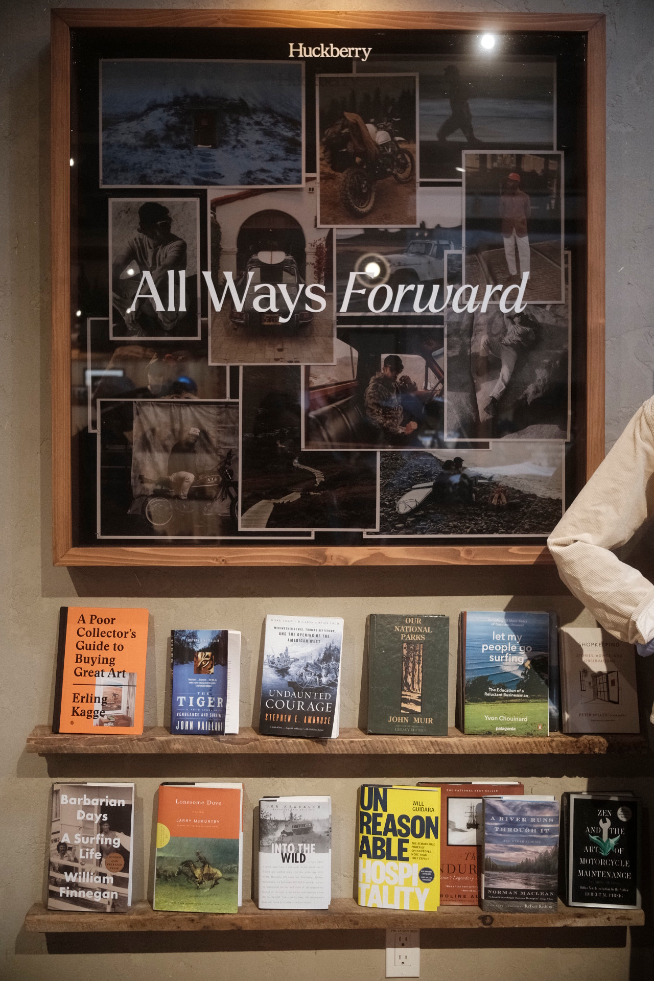 A framed collage of outdoor photographs above wooden shelves of books, a corduroy sleeve at the edge of the frame.