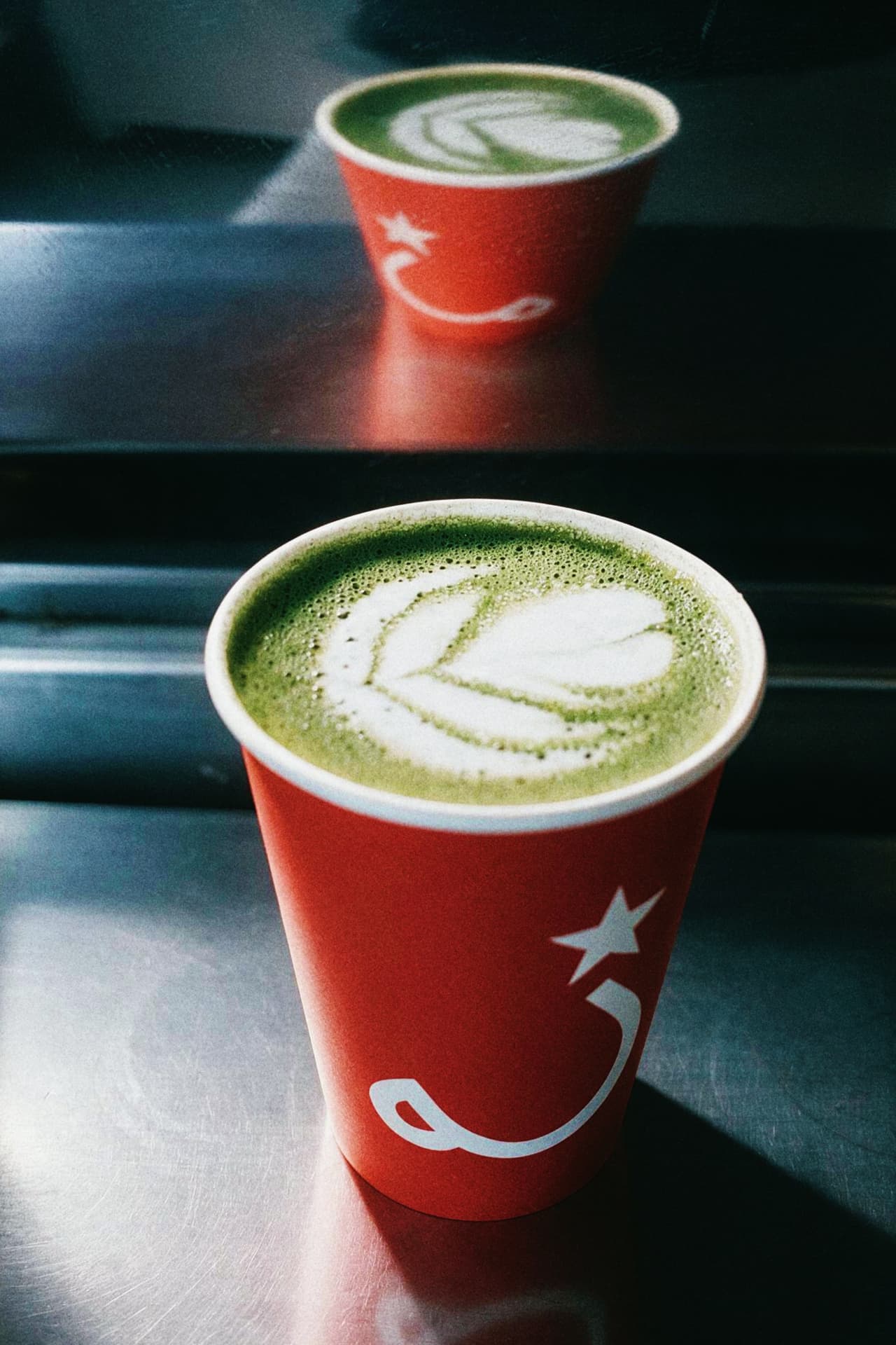Two matcha lattes with leaf latte art in coral paper cups with a white star logo, on a sunlit steel counter.
