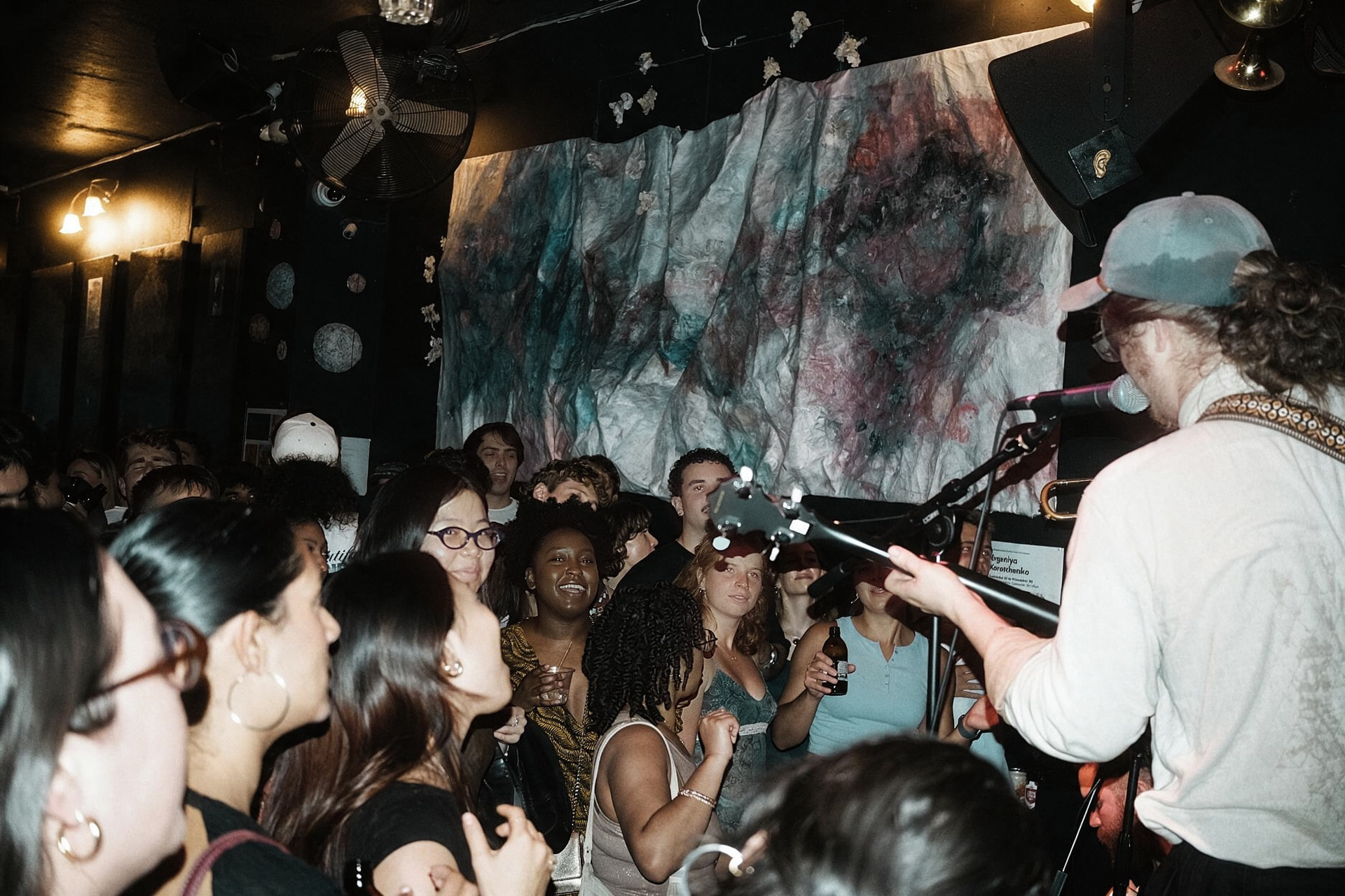 A crowded small venue at night, faces lit by flash, smiling toward a guitarist in a cap singing into a microphone.